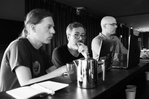 A dithered, retro-inspired photo of Martin Kleppmann, Peter van Hardenberg, and Orion Henry clustered around laptops, with looks of intense concentration on their faces.